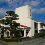 白い外壁の介護施設の建物外観。庭には松の木が植えられている。